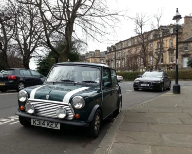 A 1992 green Classic Mini Cooper with white stripes and UK prefix registration K314 KEX, parked on a city street lined with historic sandstone buildings.