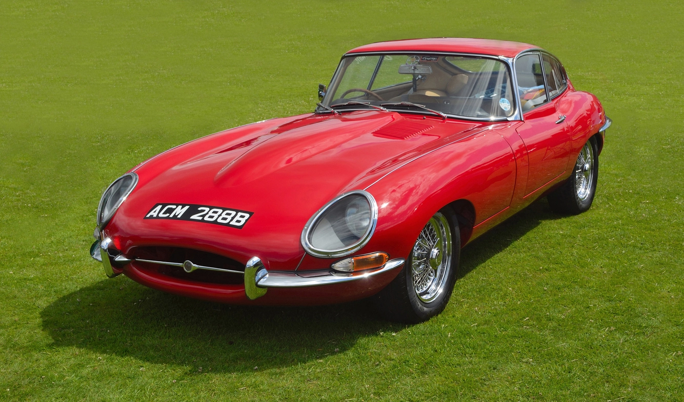 A 1964 red Jaguar E-Type with UK suffix registration ACM 288B, parked on grass, featuring chrome wire wheels and classic curved bodywork.