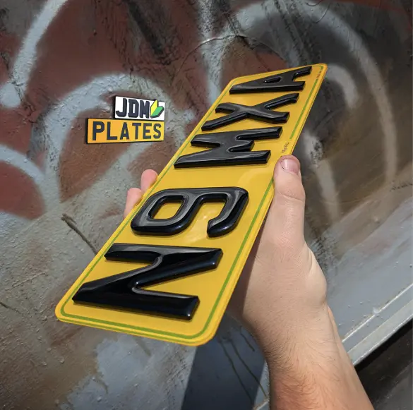 Hand holding a yellow 4D acrylic number plate with raised black characters, against a graffiti wall background, featuring the JDM Plates logo.