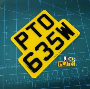Square-shaped 3D gel number plate in yellow with black characters reading "PTO 635W", produced by JDM Plates, placed on a green cutting mat alongside a small JDM Plates logo sticker.