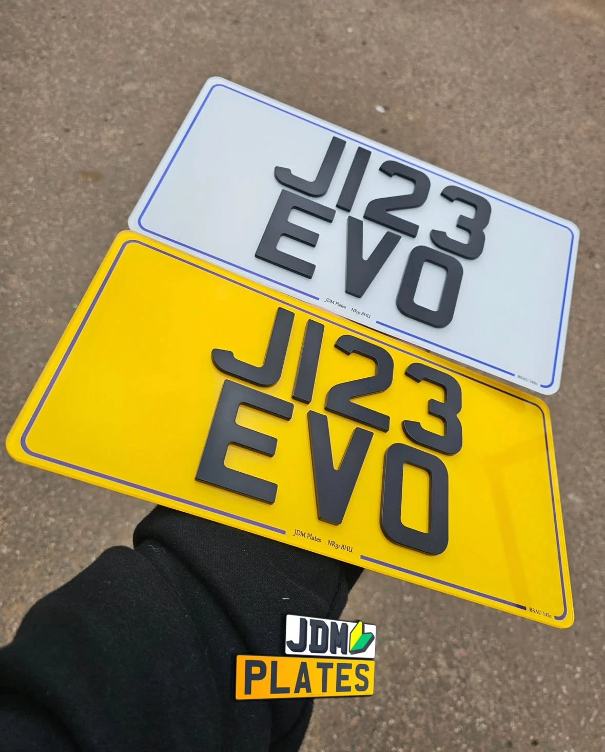 A pair of 4D number plates with the registration "J123 EVO", one in white for the front and one in yellow for the rear, both featuring laser-cut black acrylic characters. Held in hand against a tarmac background with a JDM Plates logo sticker at the bottom.