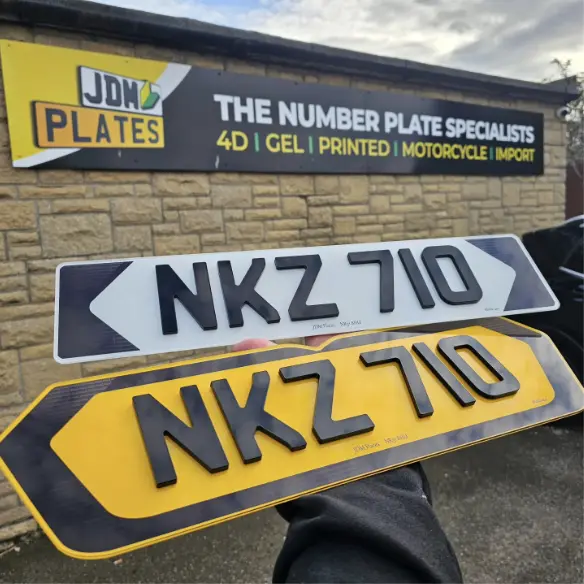 Pair of custom 4D number plates, one white front plate and one yellow rear plate with raised black acrylic characters, held in front of the JDM Plates shop sign that reads "The Number Plate Specialists – 4D, Gel, Printed, Motorcycle, Import".