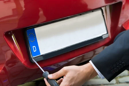 Person replacing a damaged car number plate with a screwdriver, demonstrating how to fit a new replacement plate.