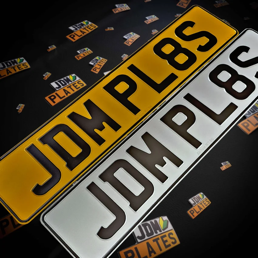A pair of yellow and white pressed metal number plate showcasing the registration number 'JDM PL8S'.