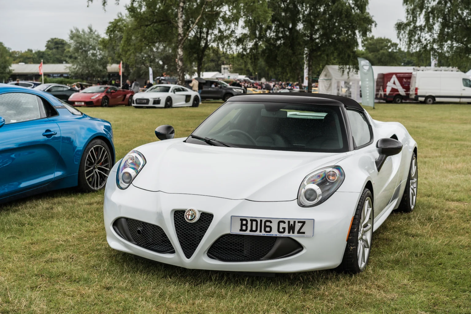 White Alfa Romeo 4C sports car on display at an outdoor car show, surrounded by other high-performance vehicles.